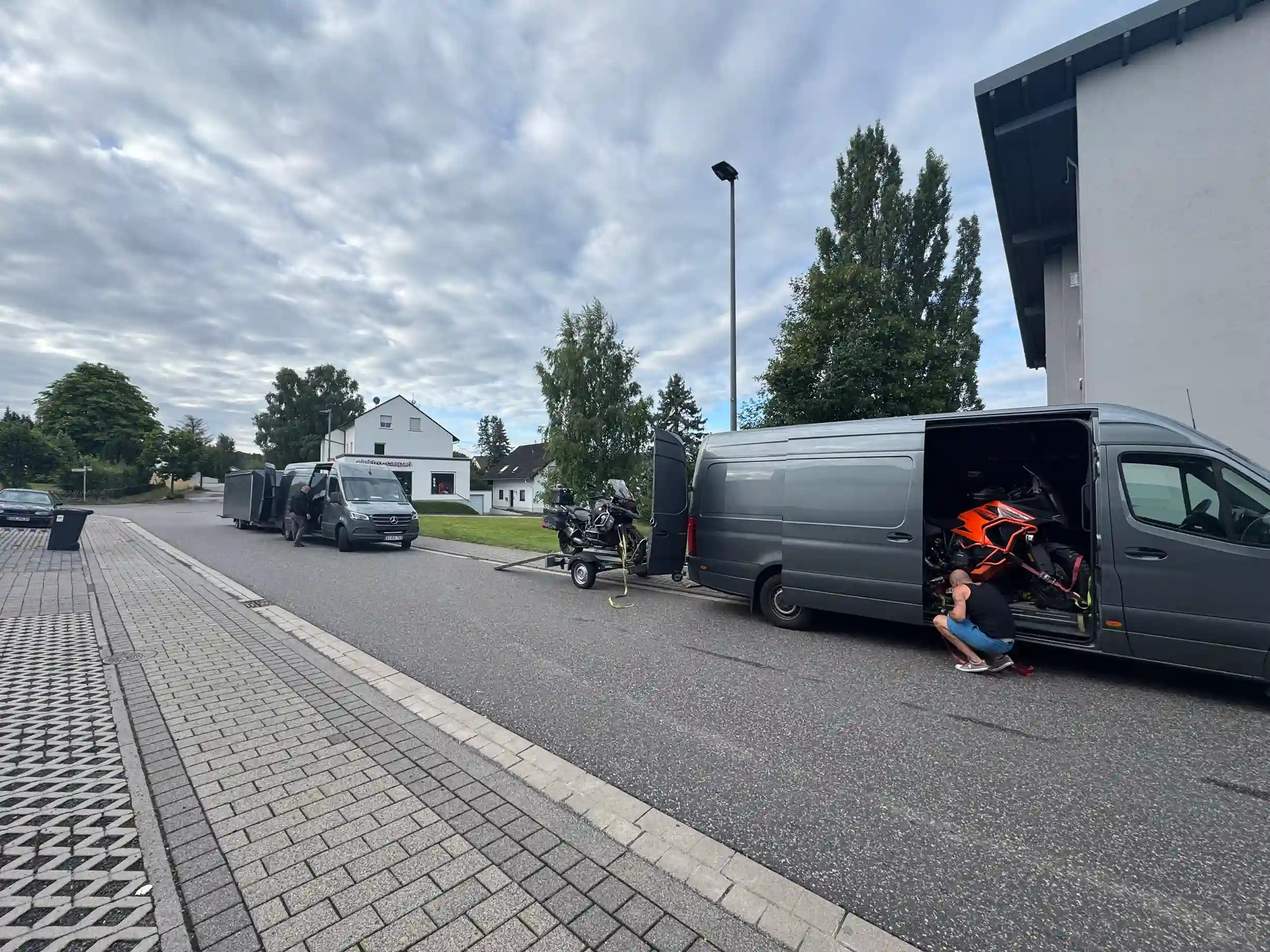 Zwei Kleintransporter und Anhänger stehen neben Häusern, Motorräder sind bereits verladen.