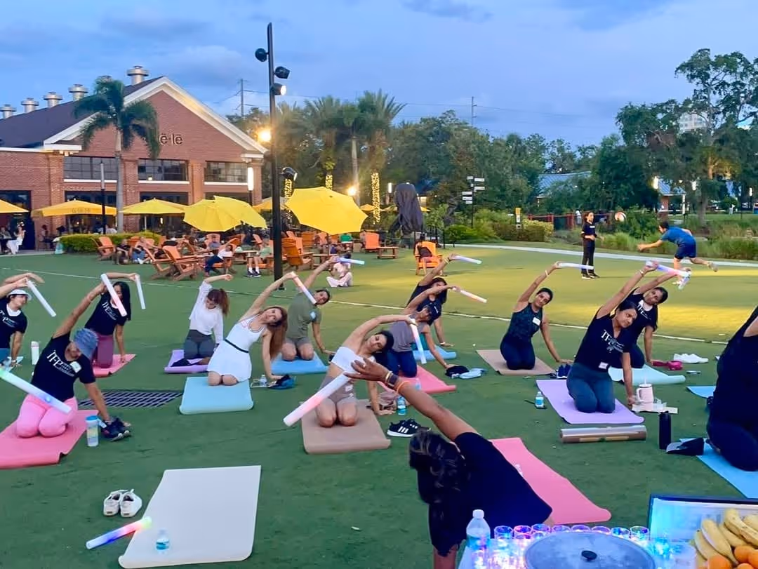 Group of people practicing outdoor yoga on mats, holding foam sticks while stretching sideways on a grassy area during evening.