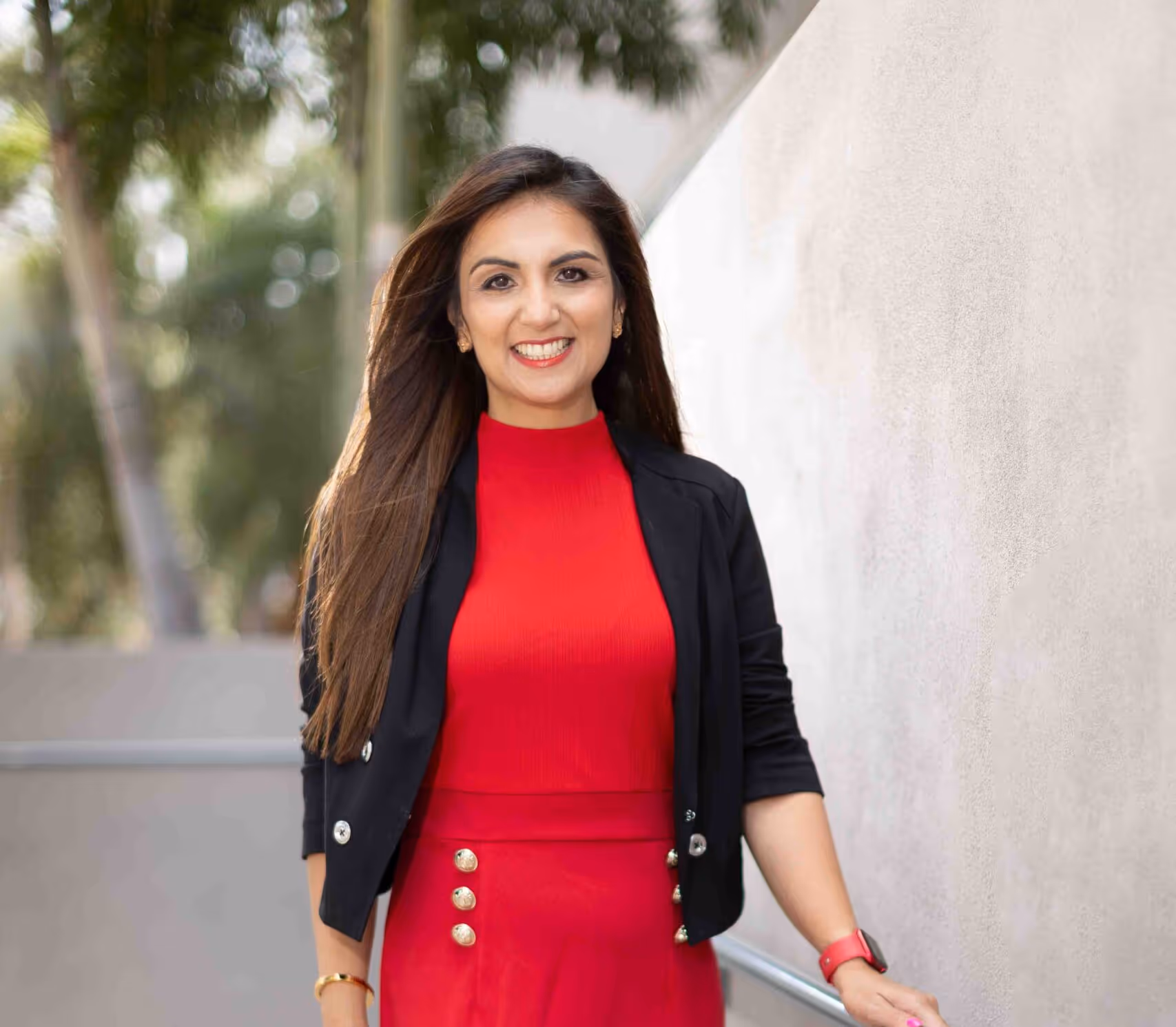 Smiling woman with long hair wearing a red dress and black blazer, standing outdoors near a light-colored wall.