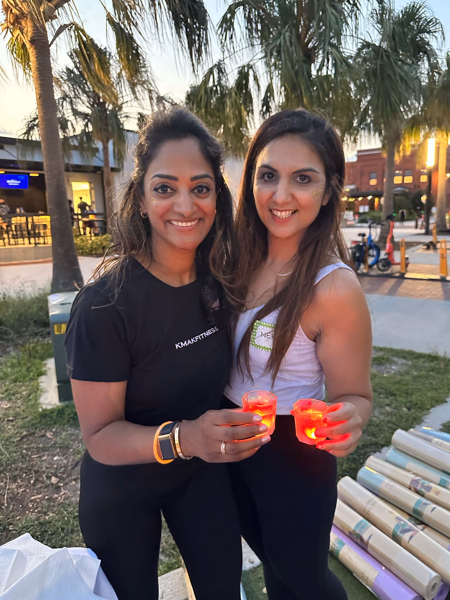 Two women smiling outdoors at dusk holding small glowing red lanterns, standing near palm trees.