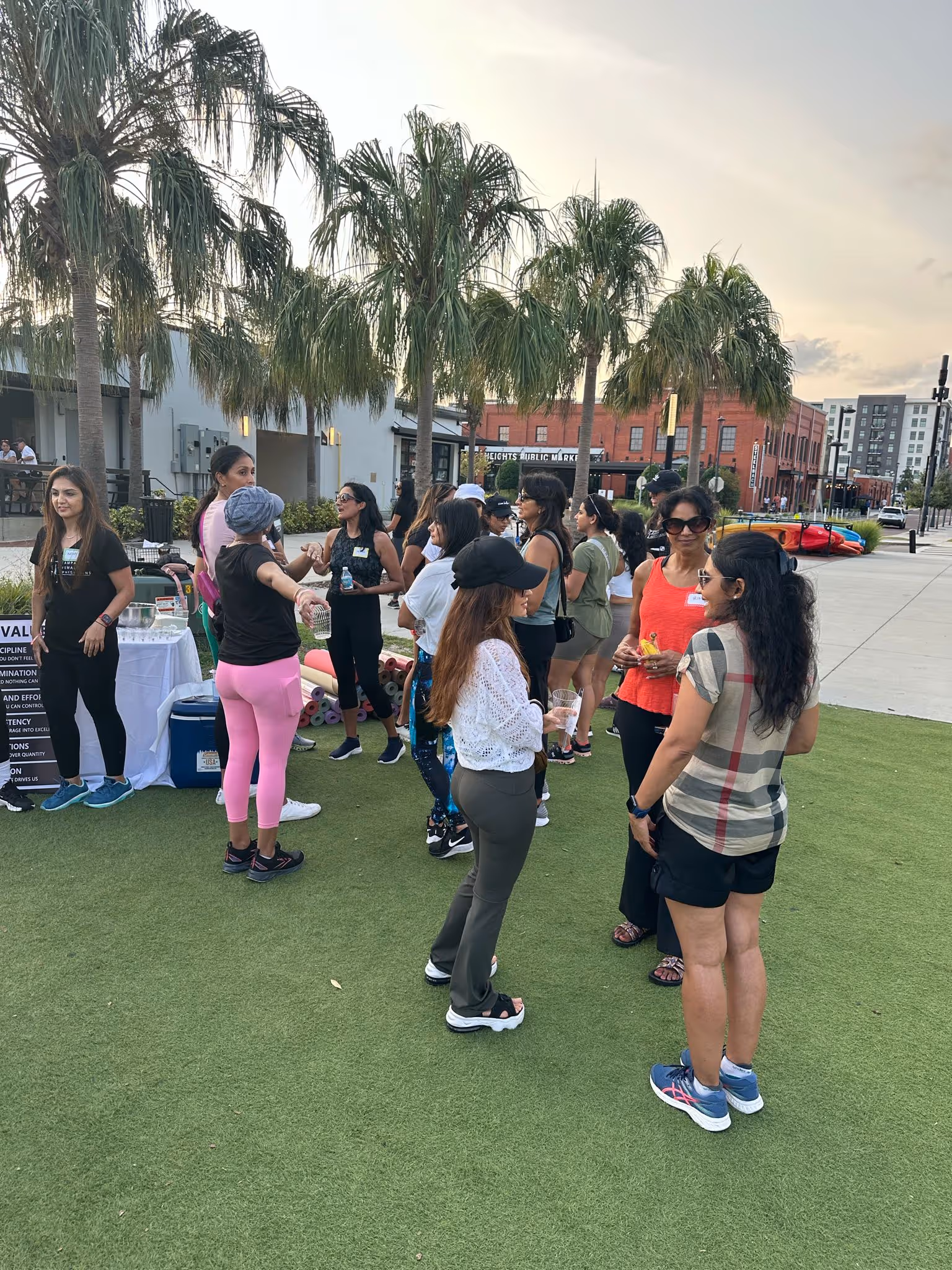 Group of people socializing outdoors on artificial grass near palm trees and a building at sunset.