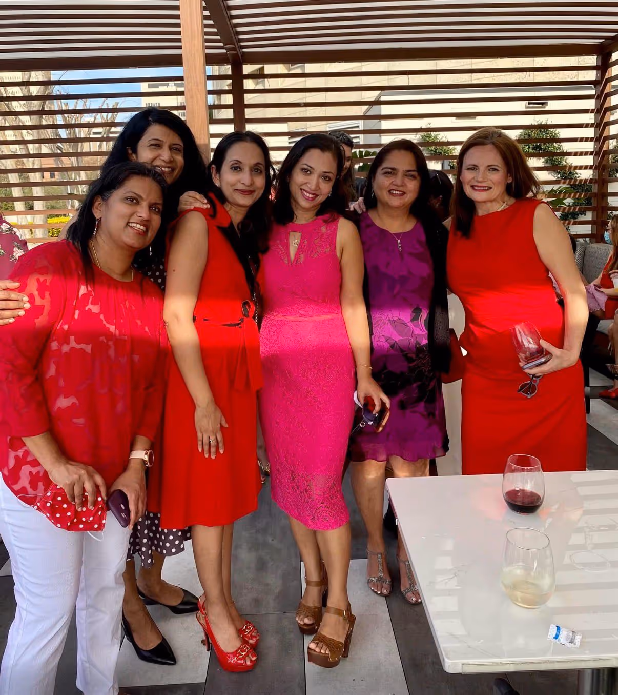 Group of six women smiling and posing together under a pergola, dressed in colorful dresses, with two wine glasses on a nearby table.