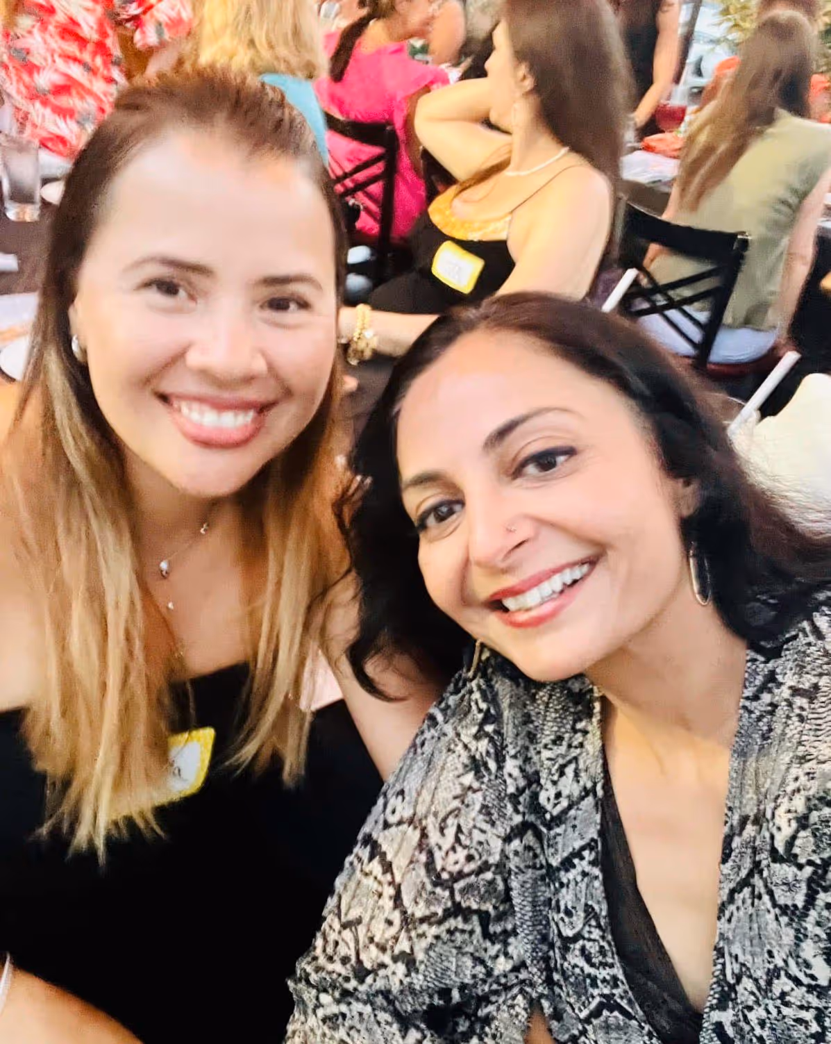 Two women smiling and posing for a close-up selfie at a social gathering with people seated in the background.