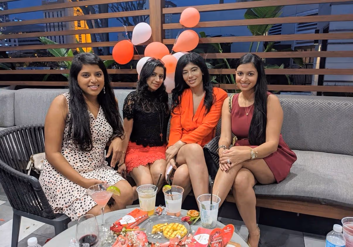 Four women sitting on a gray sofa around a table with drinks and food, with red and white balloons in the background.