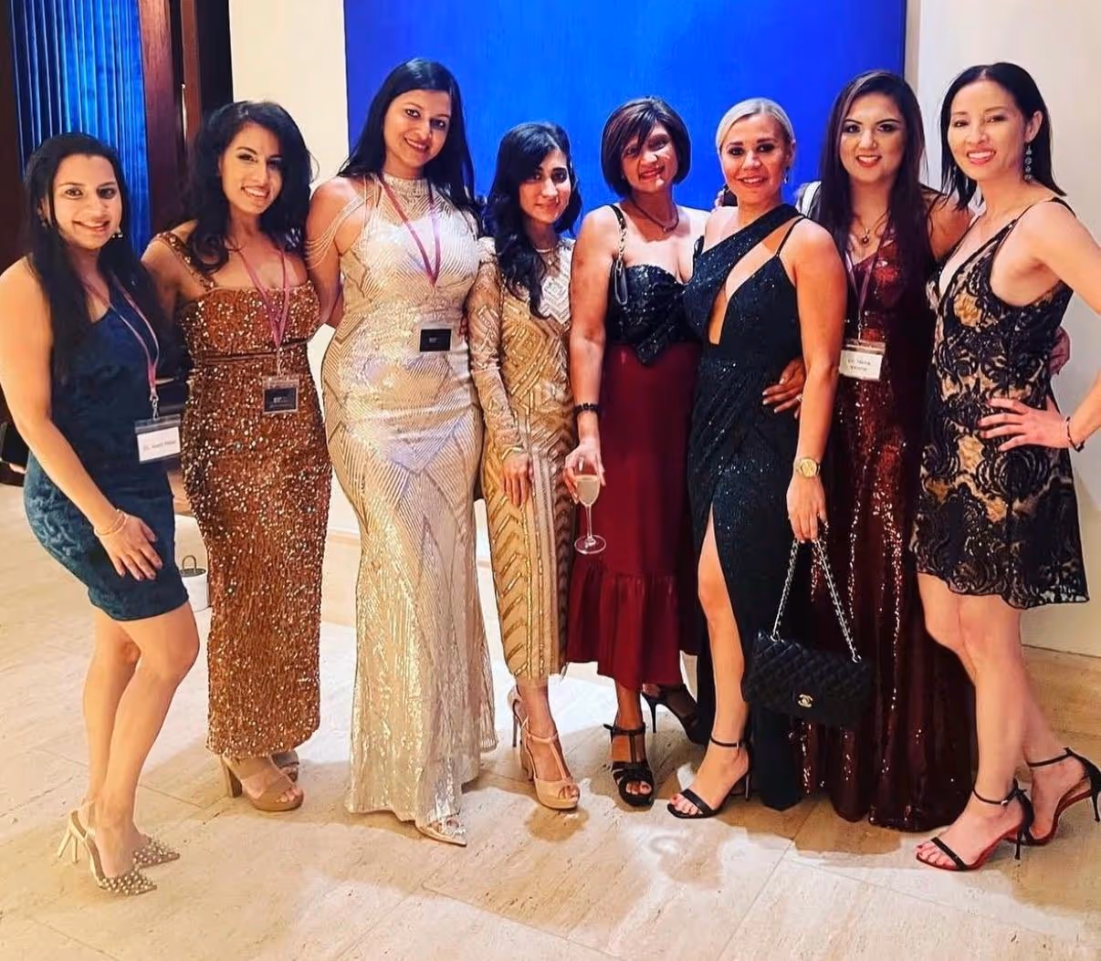 Group of eight women dressed in elegant evening gowns posing indoors, some wearing name badges and high heels.