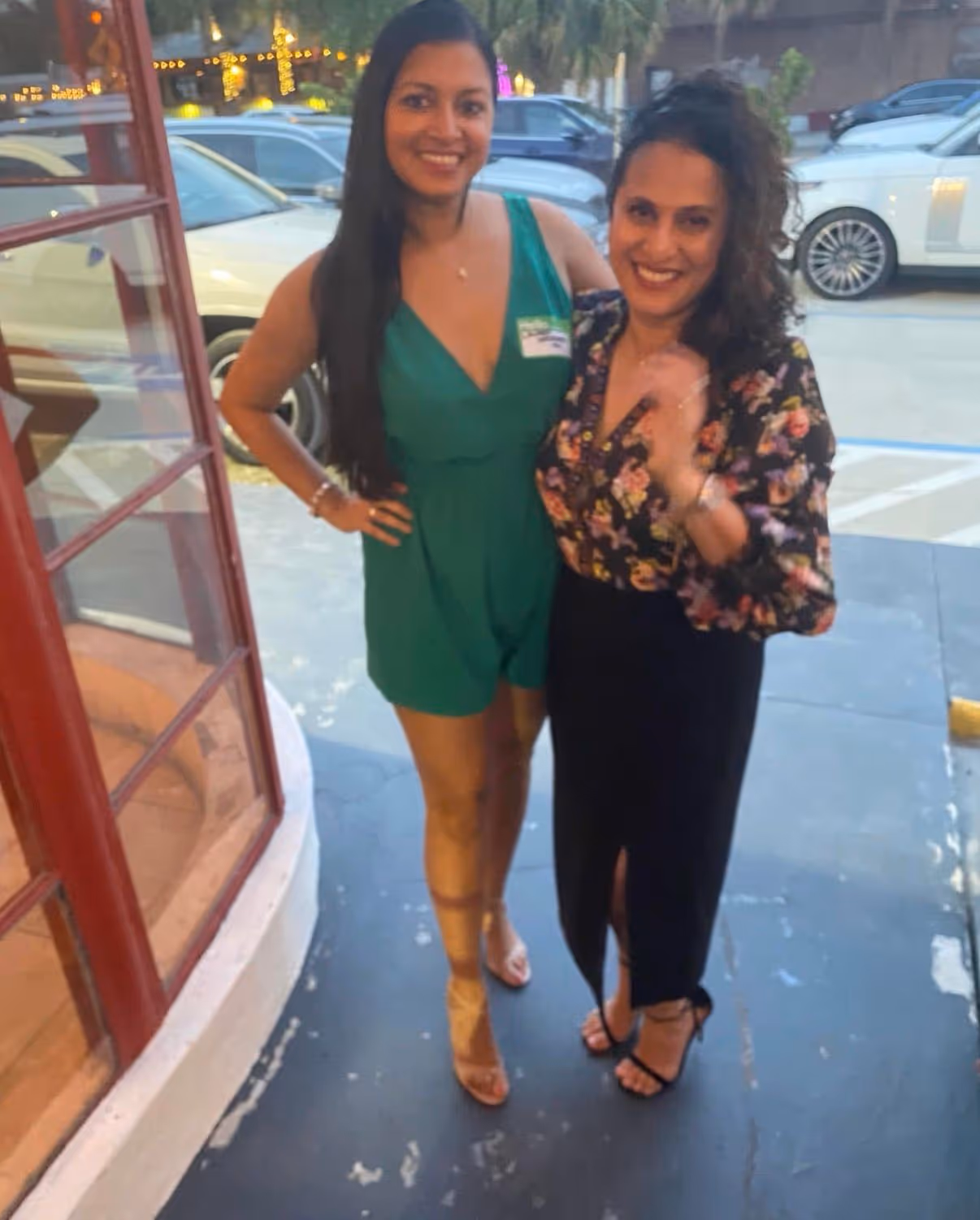 Two women smiling and posing together outside by a building with large windows and parked cars in the background.