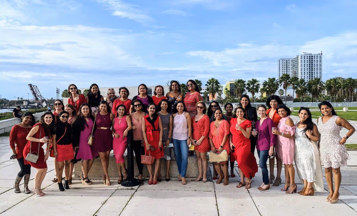 A large group of women dressed mostly in red and pink posing outdoors on a sunny day with palm trees and buildings in the background.