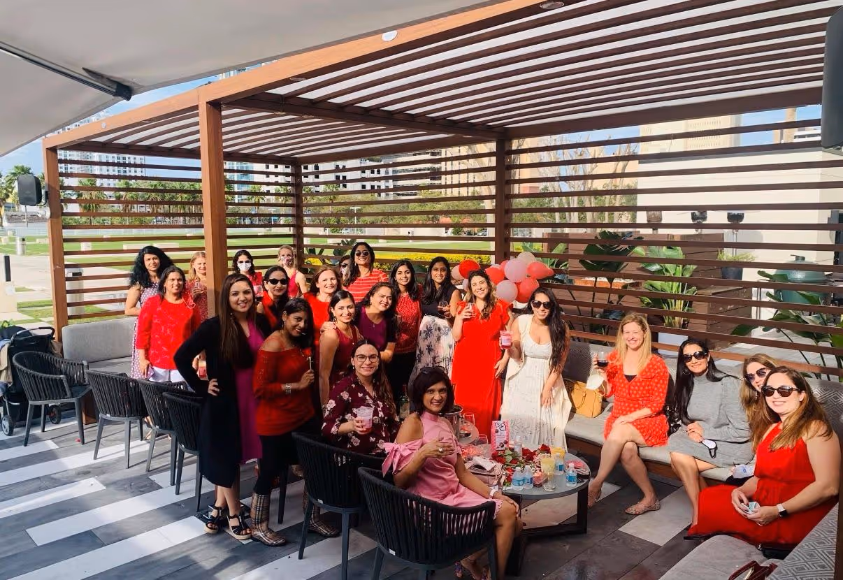 Group of women gathered under a wooden pergola, some sitting and others standing, wearing red and pink outfits, enjoying a social event with drinks and balloons.