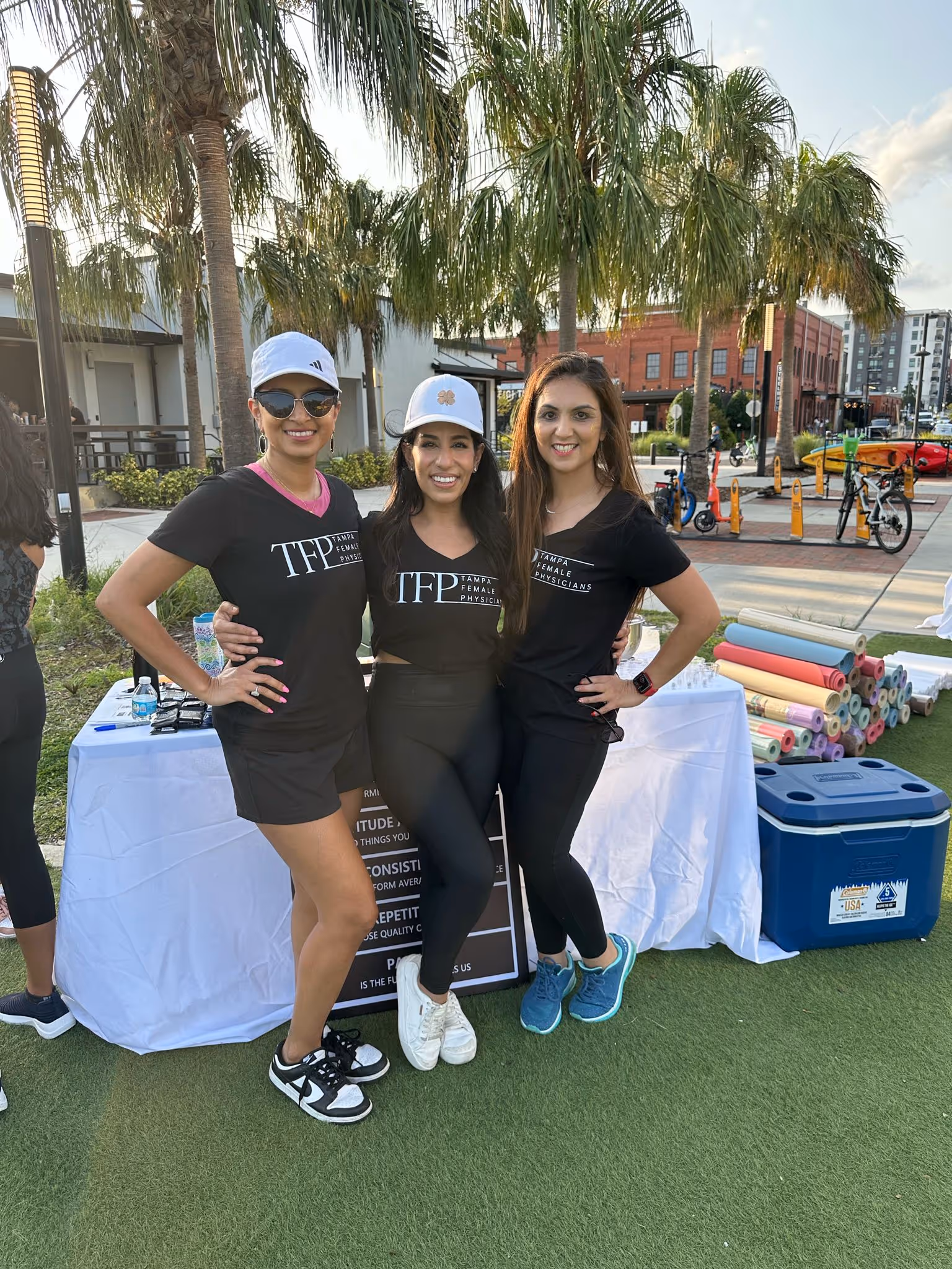Three women smiling and posing outdoors wearing black Tampa Female Physicians t-shirts with palm trees and yoga mats in the background.