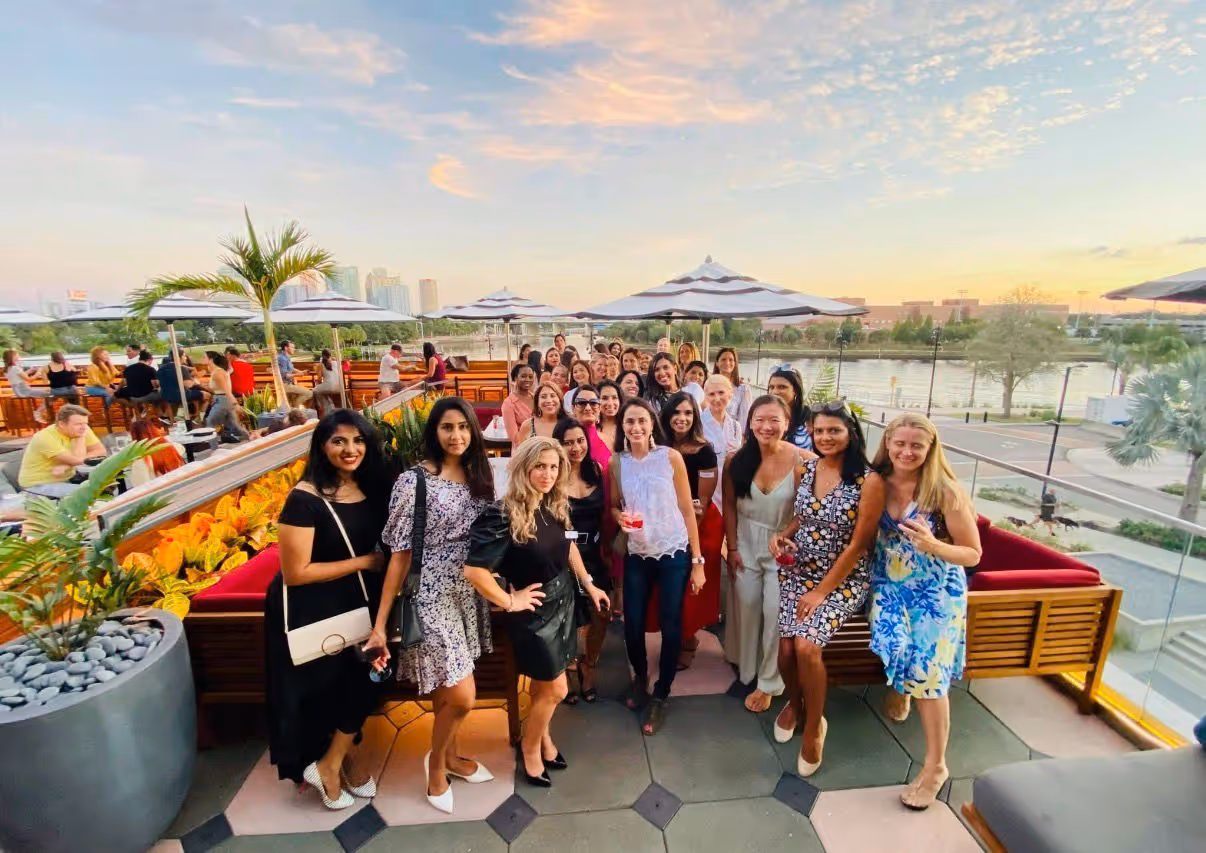 Group of women gathered and smiling on an outdoor patio with umbrellas, plants, and a river view at sunset.