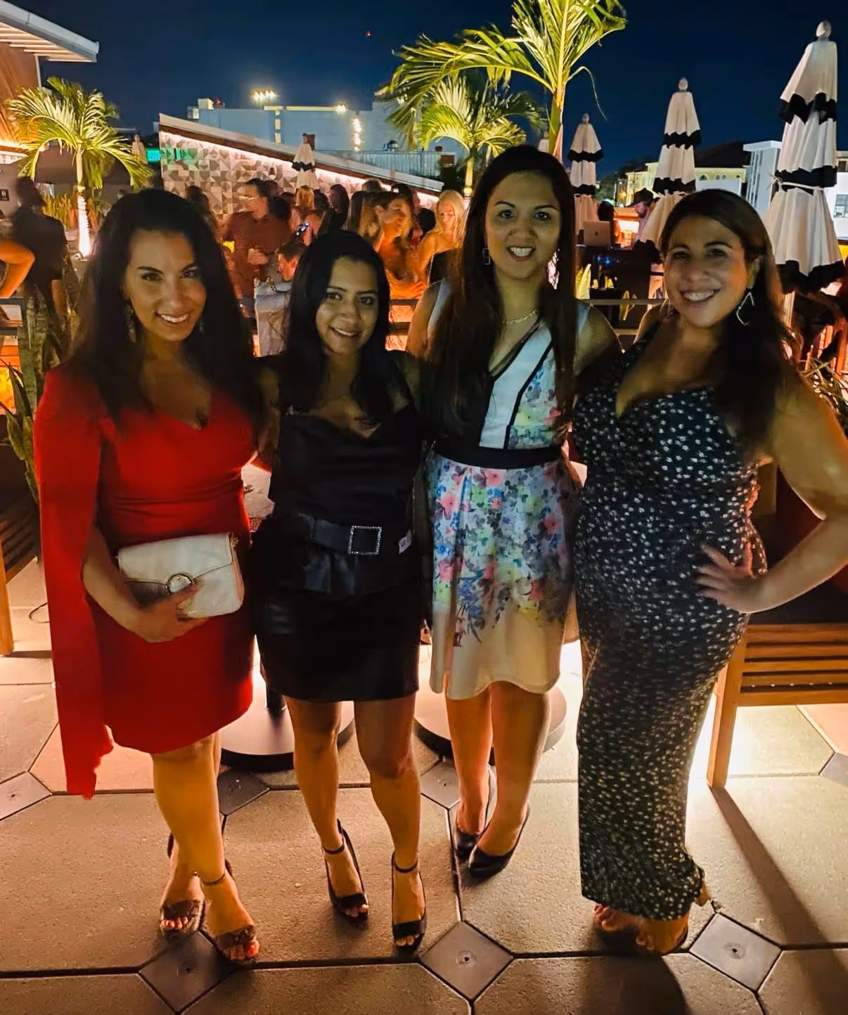 Four women dressed in evening attire posing together outdoors at night with palm trees and umbrellas in the background.