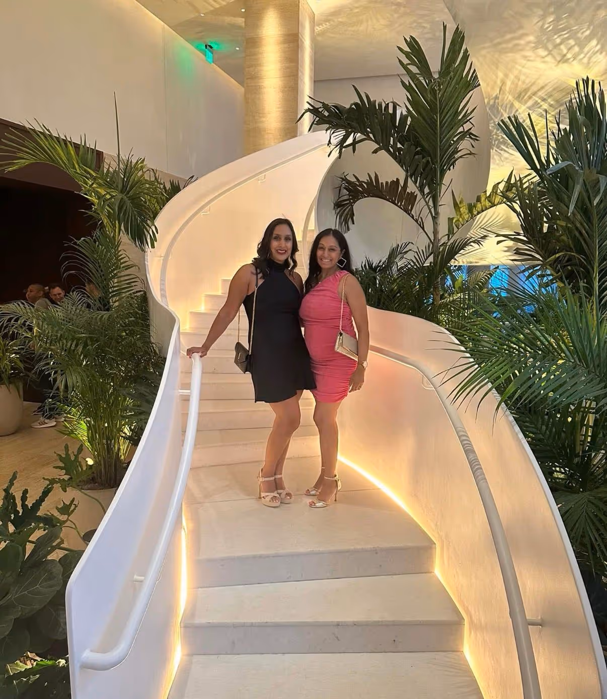 Two women standing on an illuminated white curved staircase surrounded by green plants.