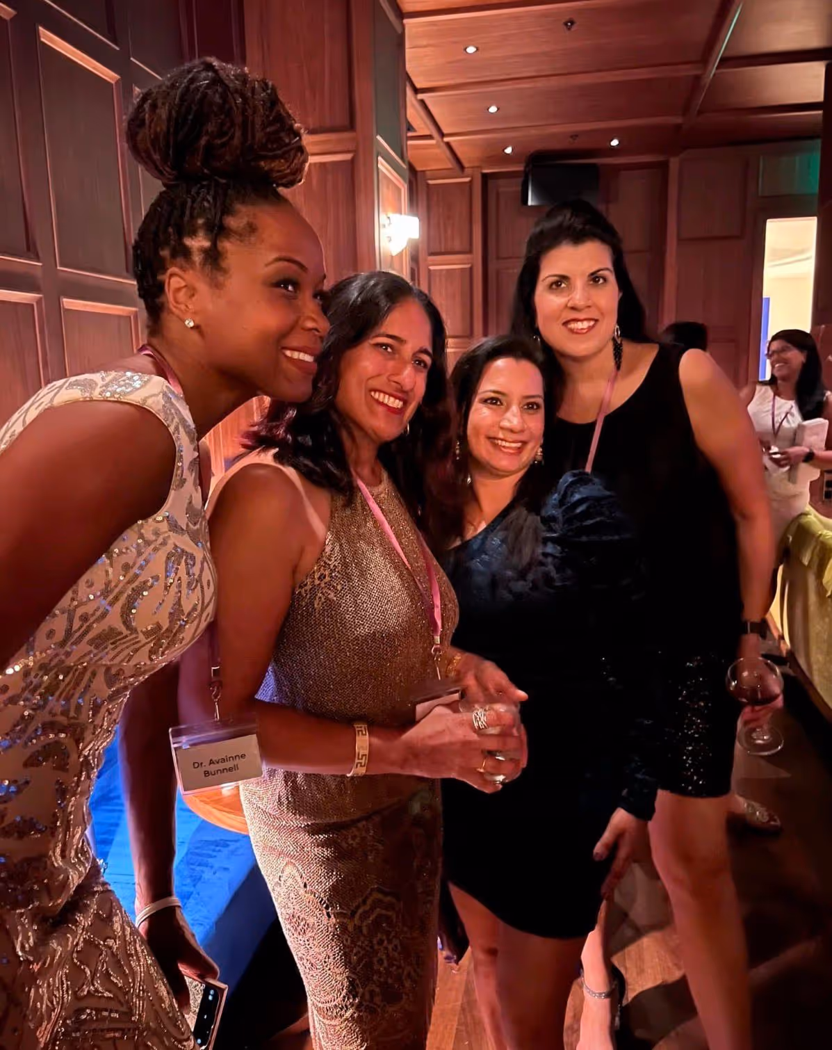 Four women dressed in formal attire smiling and posing together at an indoor event.