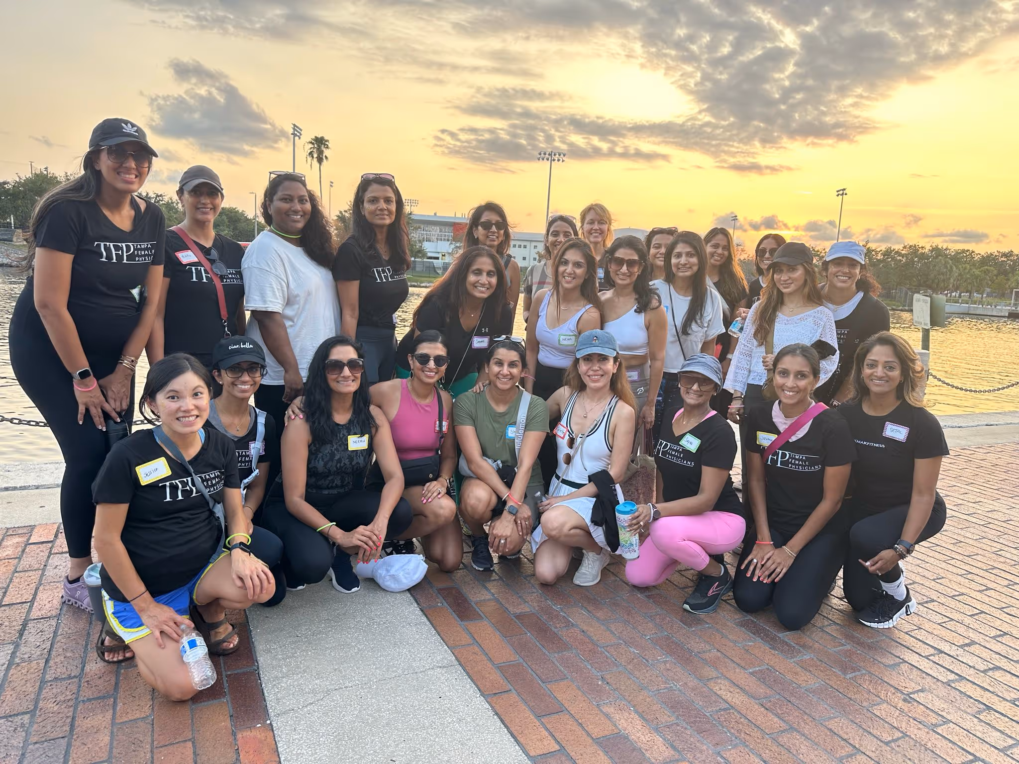 Group of diverse women smiling outdoors near water at sunset, many wearing Tampa Female Physicians shirts.