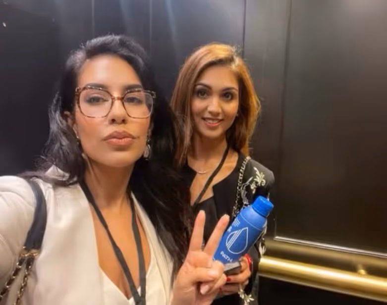 Two women posing for a selfie in an elevator, one making a peace sign and the other holding a blue water bottle.