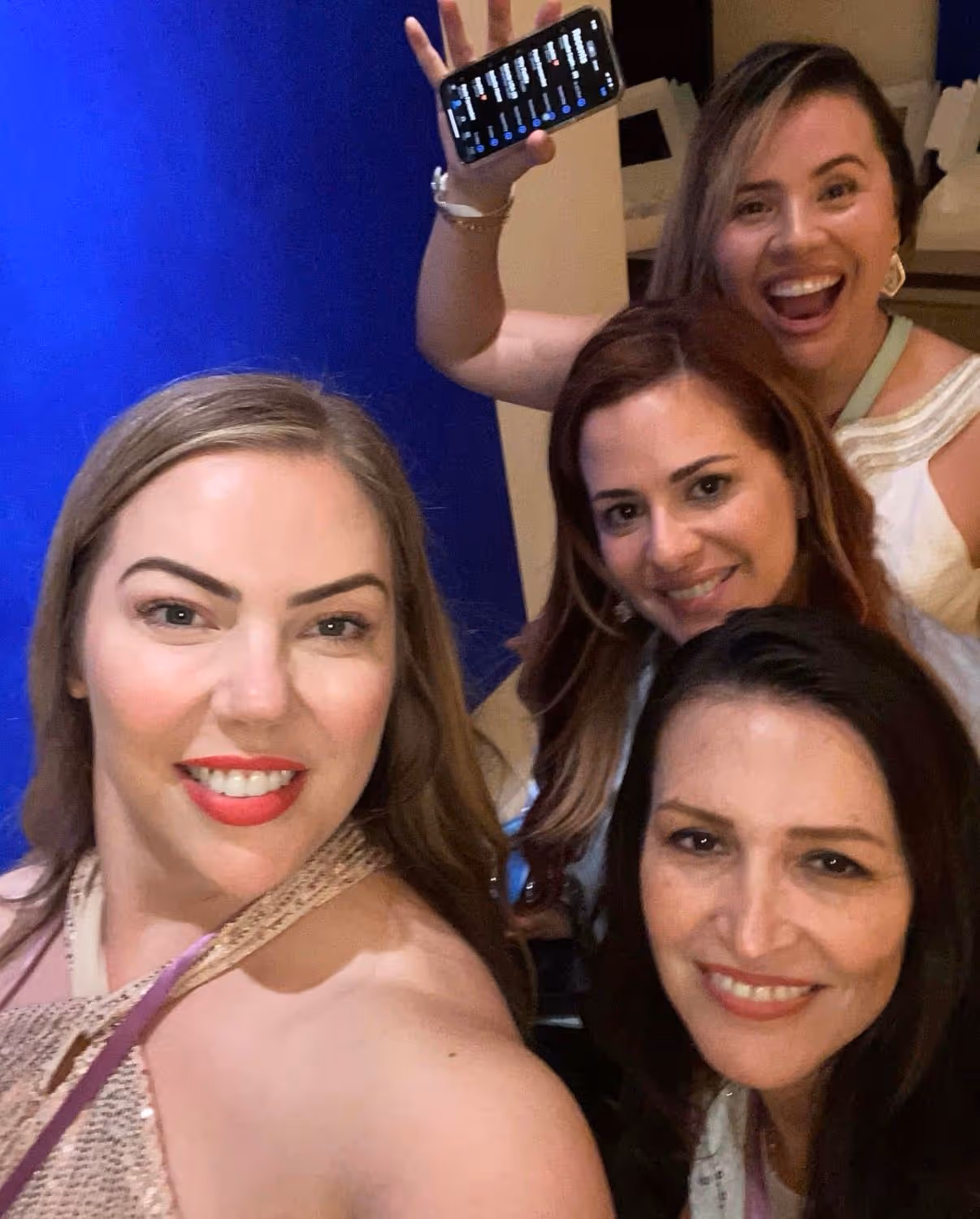 Four smiling women posing for a selfie, one holding a phone with a dark screen above her head.