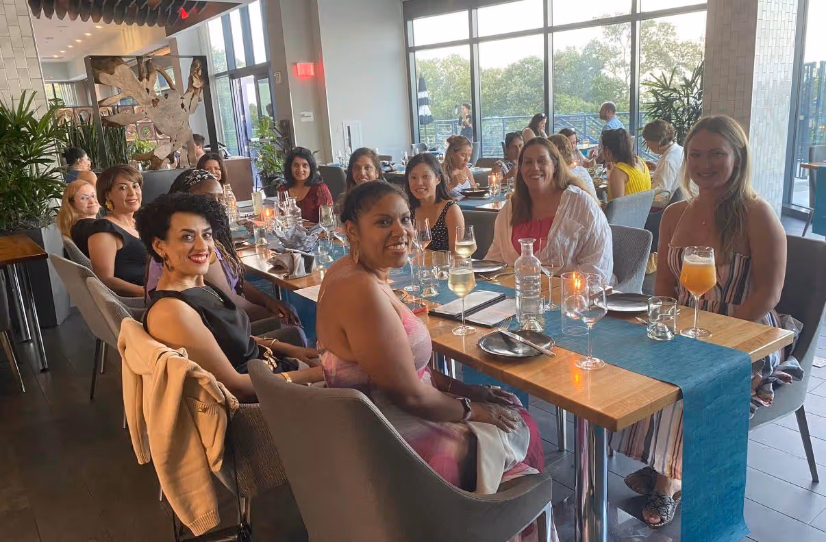 Group of women smiling and seated around a long dining table with drinks and menus in a bright, modern restaurant.
