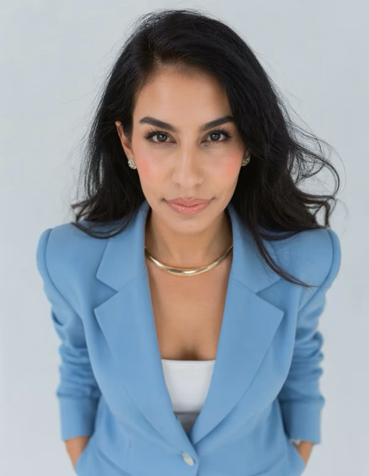 Portrait of a woman with long dark hair wearing a light blue blazer and gold necklace, looking confidently at the camera against a light background.