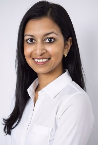 Smiling woman with long black hair wearing a white shirt against a plain light background.