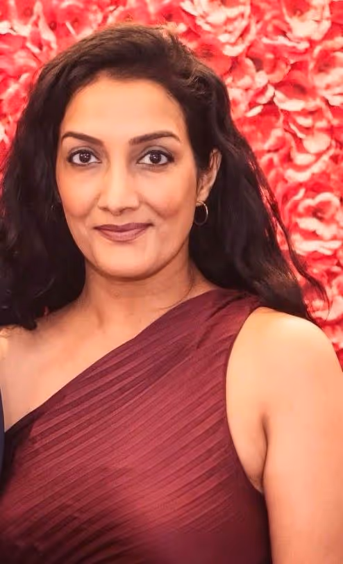 Woman with long black hair wearing a sleeveless burgundy dress, standing in front of a background of red roses.