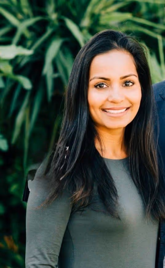 Smiling woman with long dark hair wearing a green top standing outdoors with leafy green plants in the background.