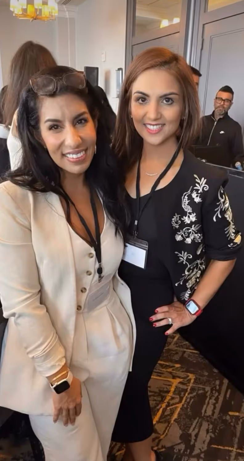 Two smiling women at an indoor event wearing name badges and smart casual attire, one in a light-colored suit and the other in a black dress with a floral jacket.