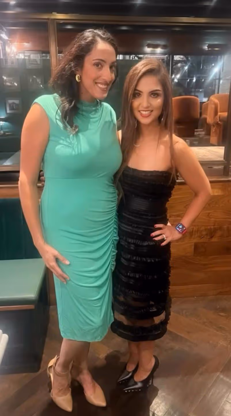 Two women smiling indoors, one wearing a green ruched dress with beige heels, the other in a black strapless dress with black heels and a smartwatch.