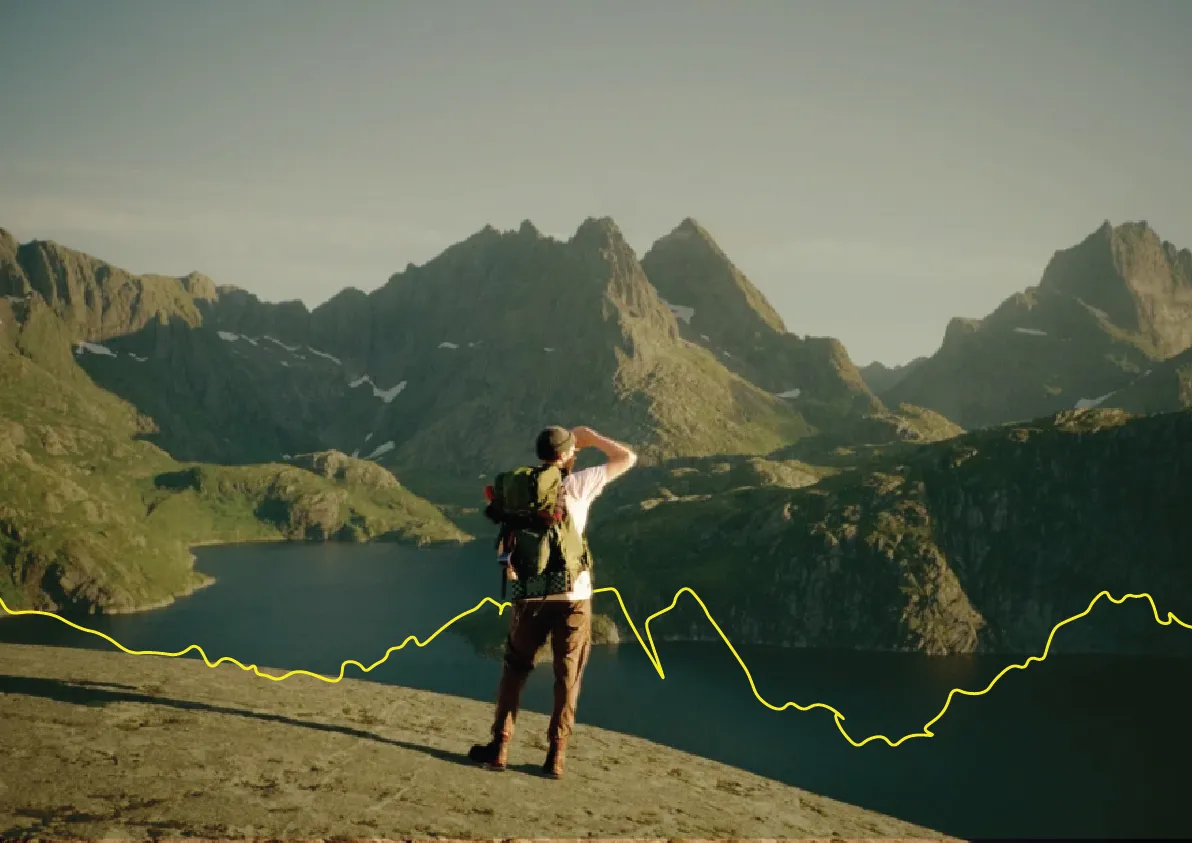 Een wandelaar staat op een rots met een rugzak en kijkt naar een berglandschap met een meer en scherpe toppen.