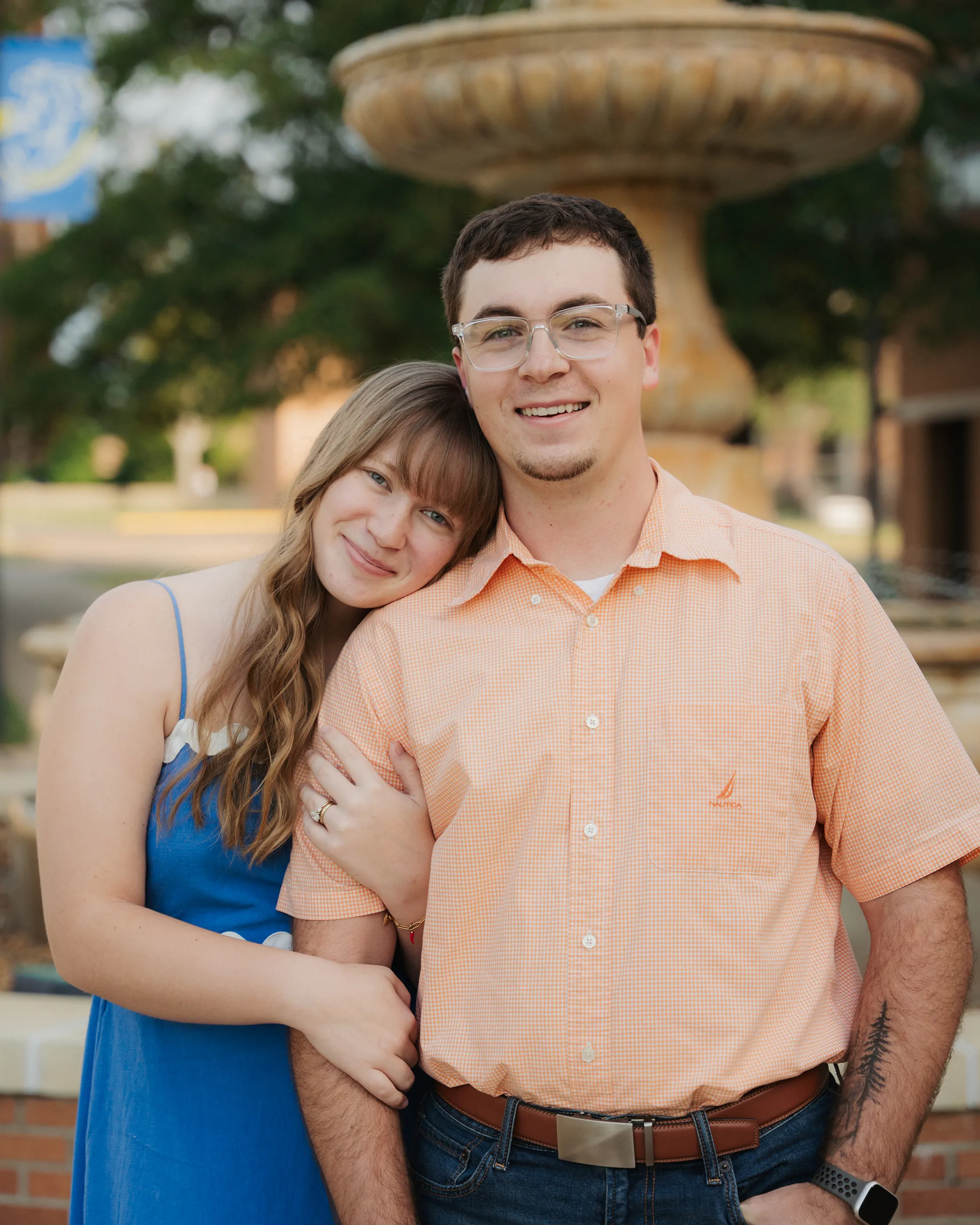 Smiling couple standing close together outside with woman resting her head on man's shoulder by a fountain.