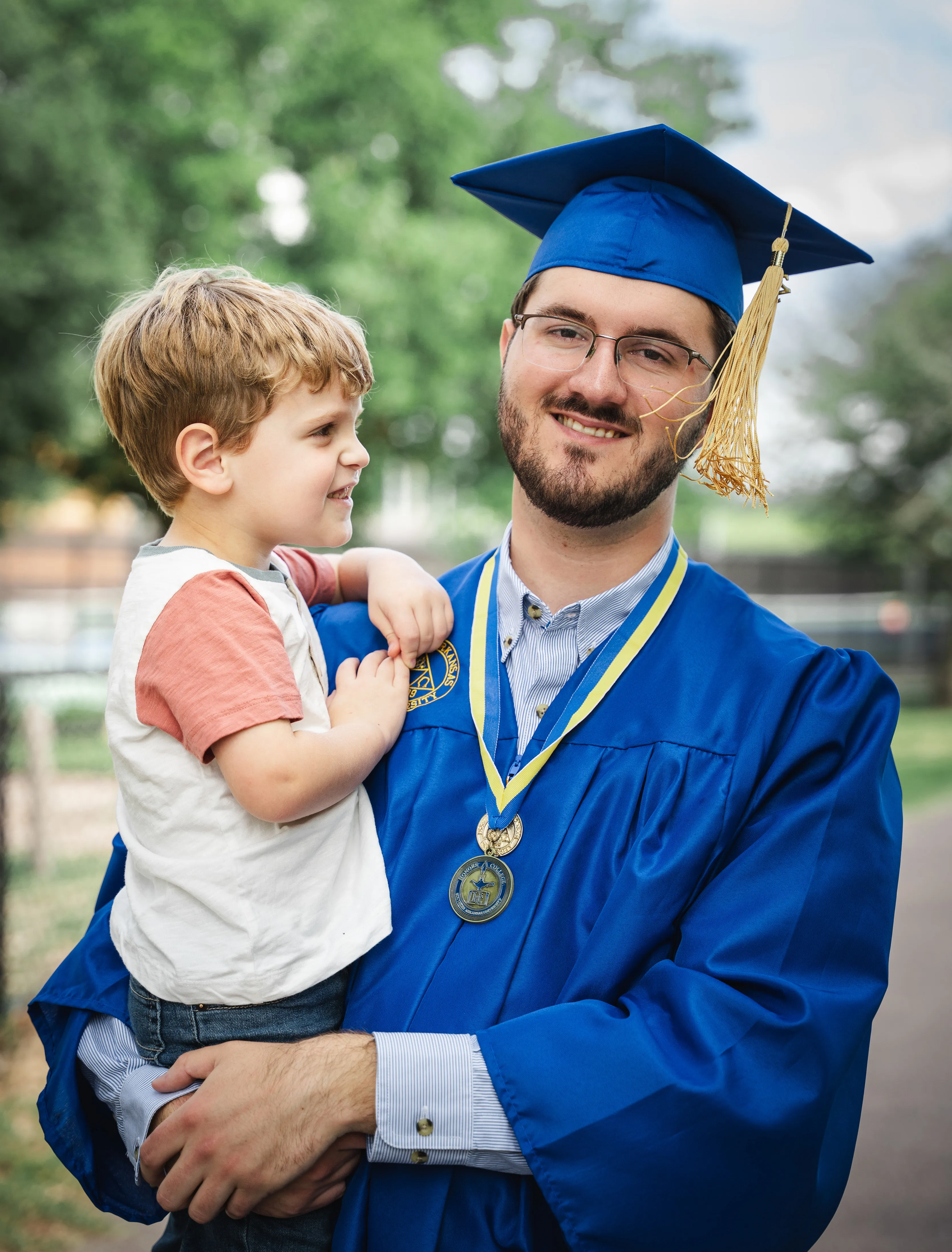 Young man in blue graduation gown and cap holding a small boy outdoors, both smiling.