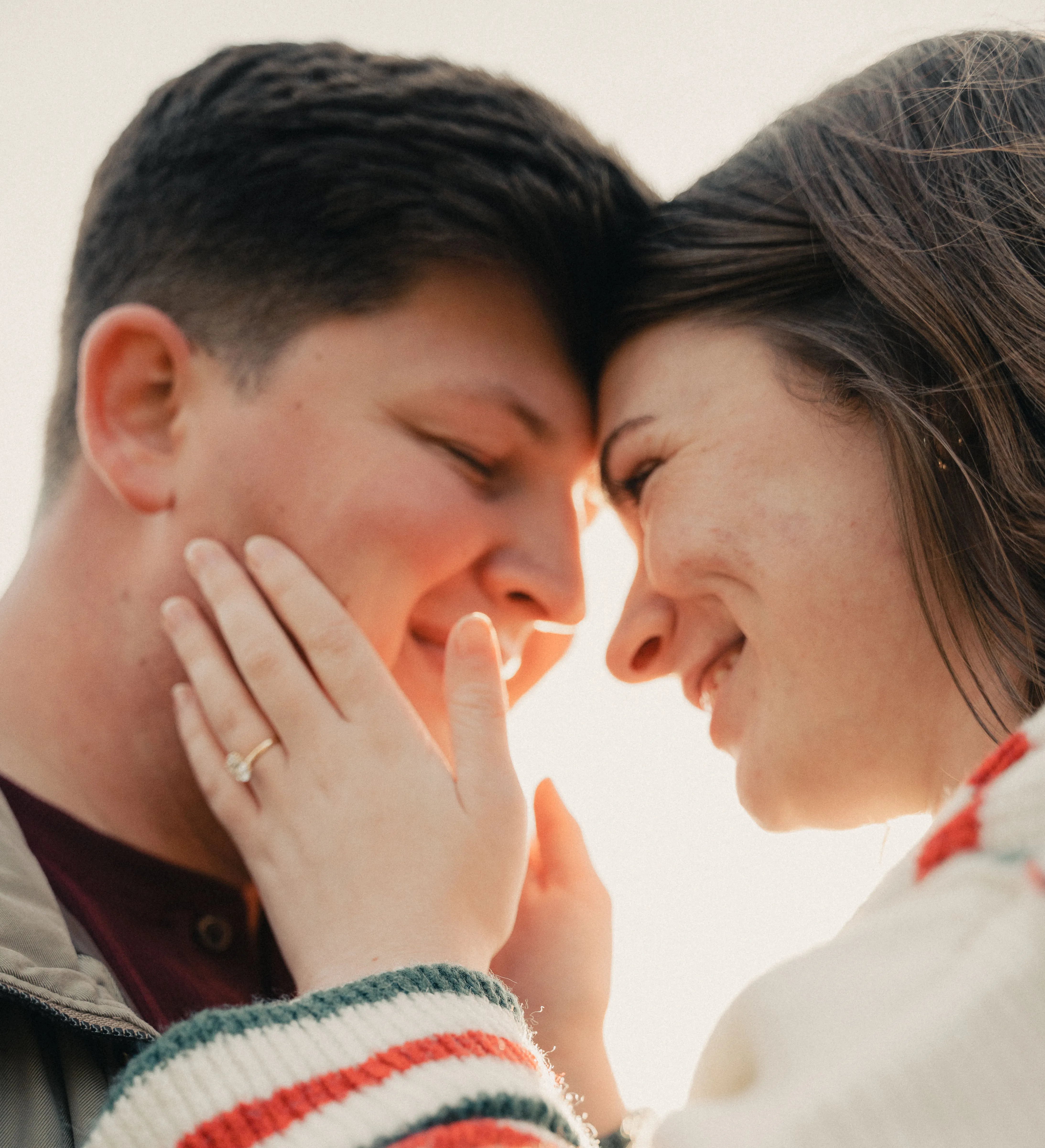 Close-up of a smiling couple touching foreheads, the woman gently holding the man's face with an engagement ring visible on her finger.