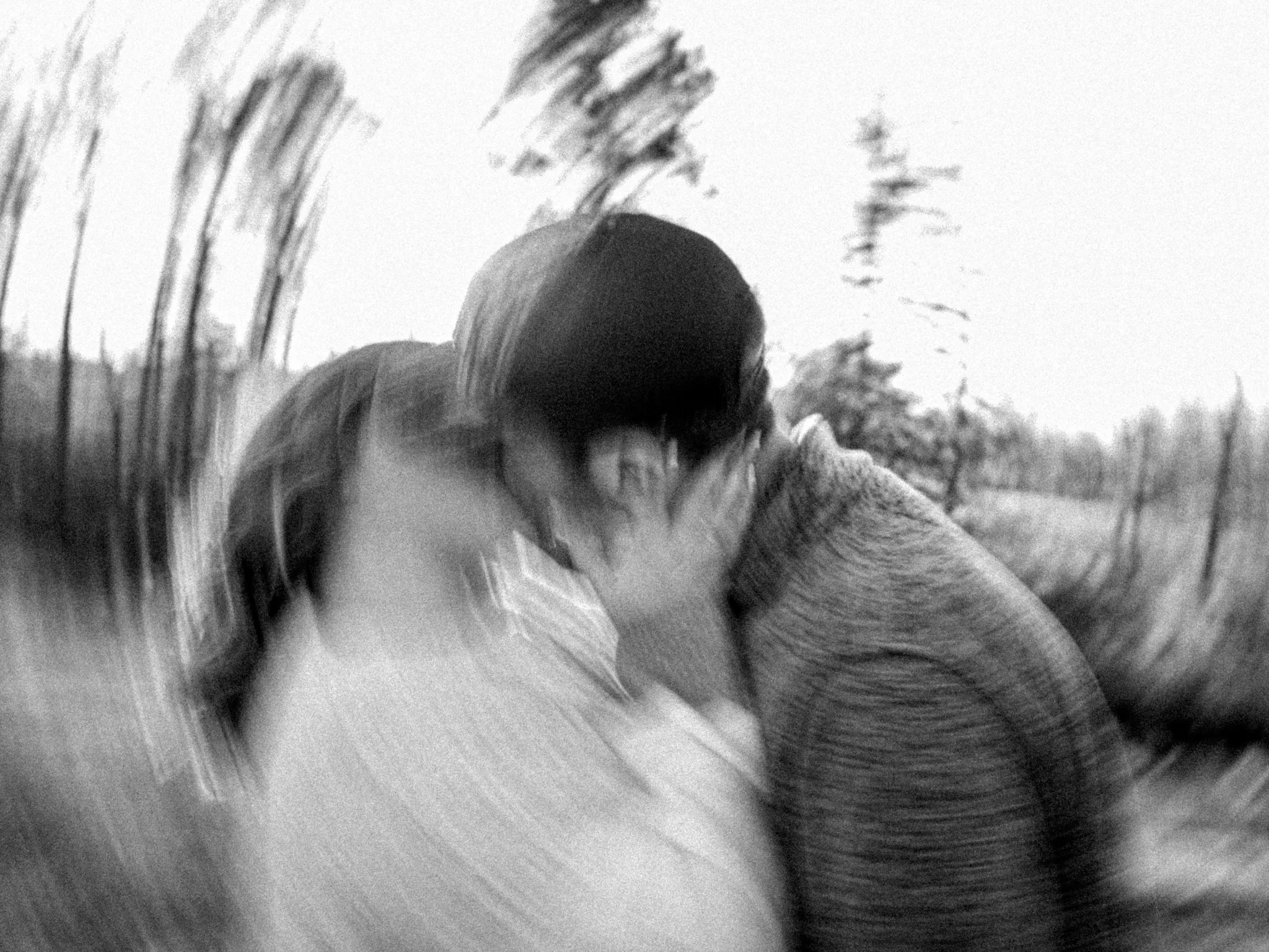 Black and white blurred photo of a couple kissing outdoors with trees in the background.