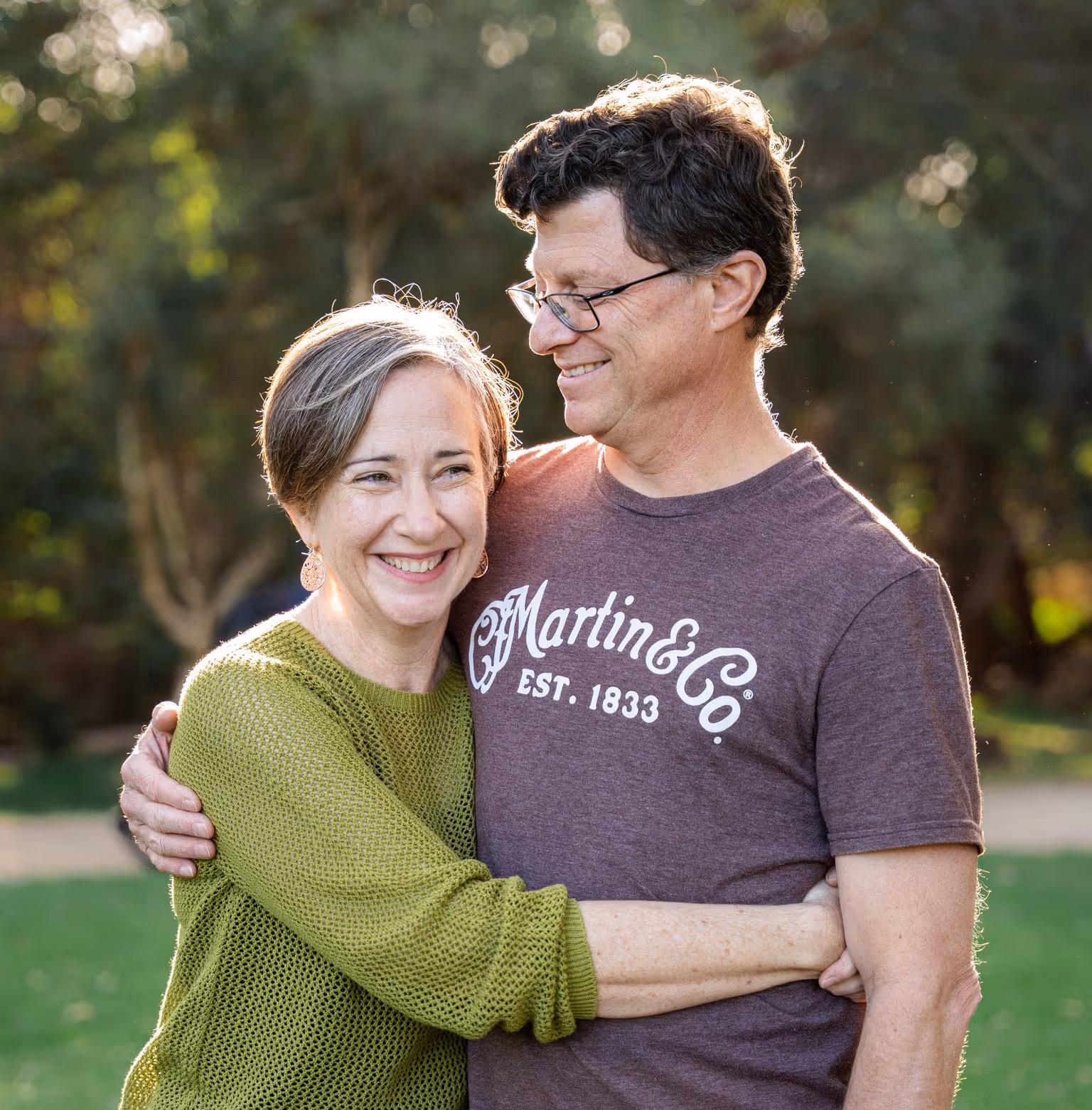 Smiling middle-aged couple hugging outdoors with greenery in the background.