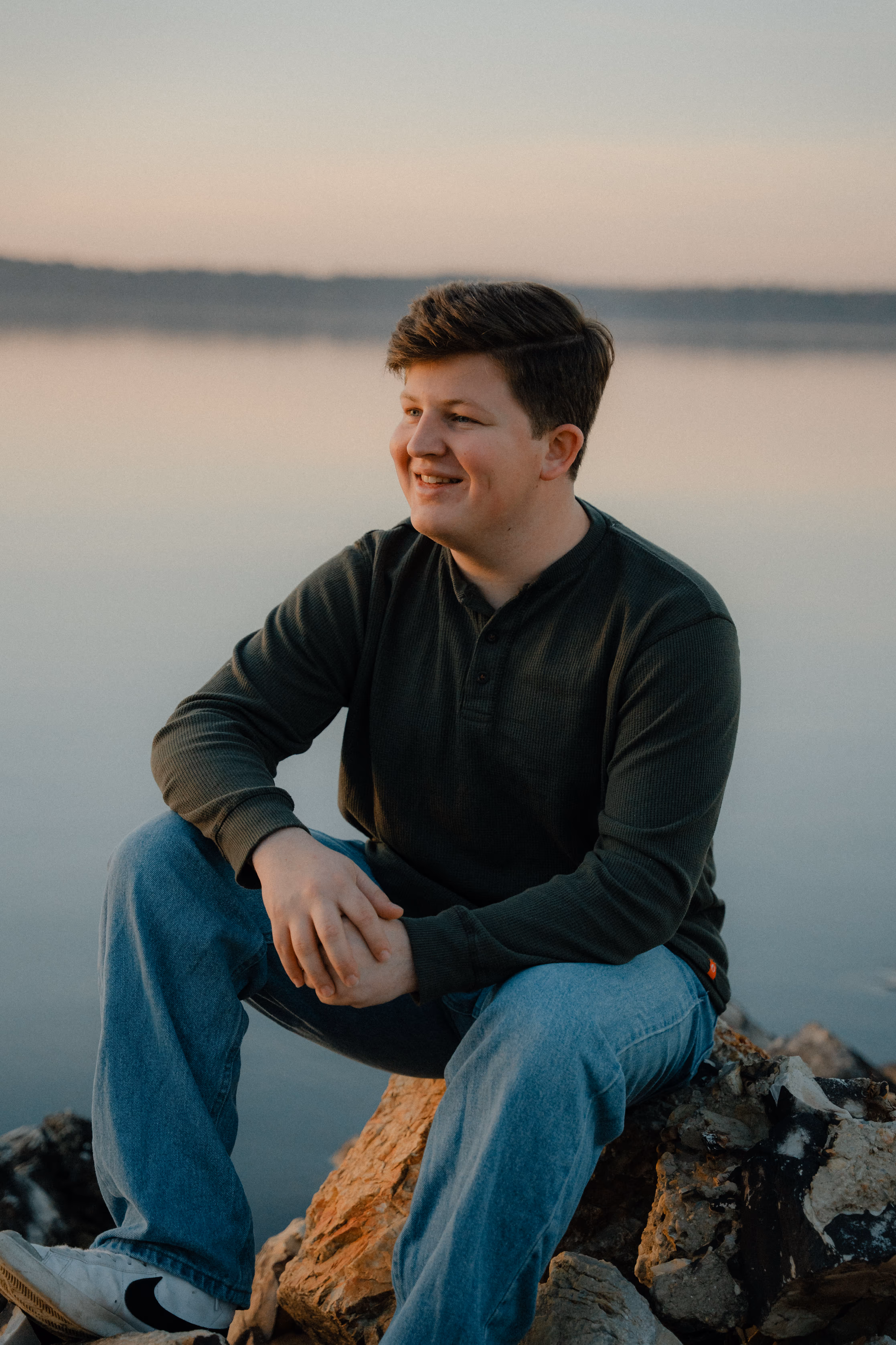 Young man smiling and sitting on rocks by calm water at sunset.