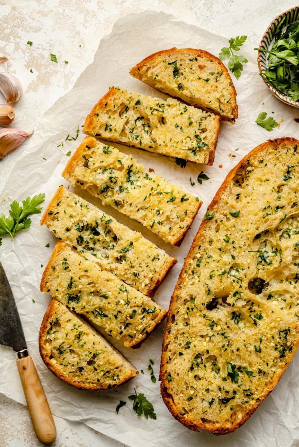 Sliced garlic bread topped with parsley on parchment paper. A knife and garlic cloves lie alongside.
