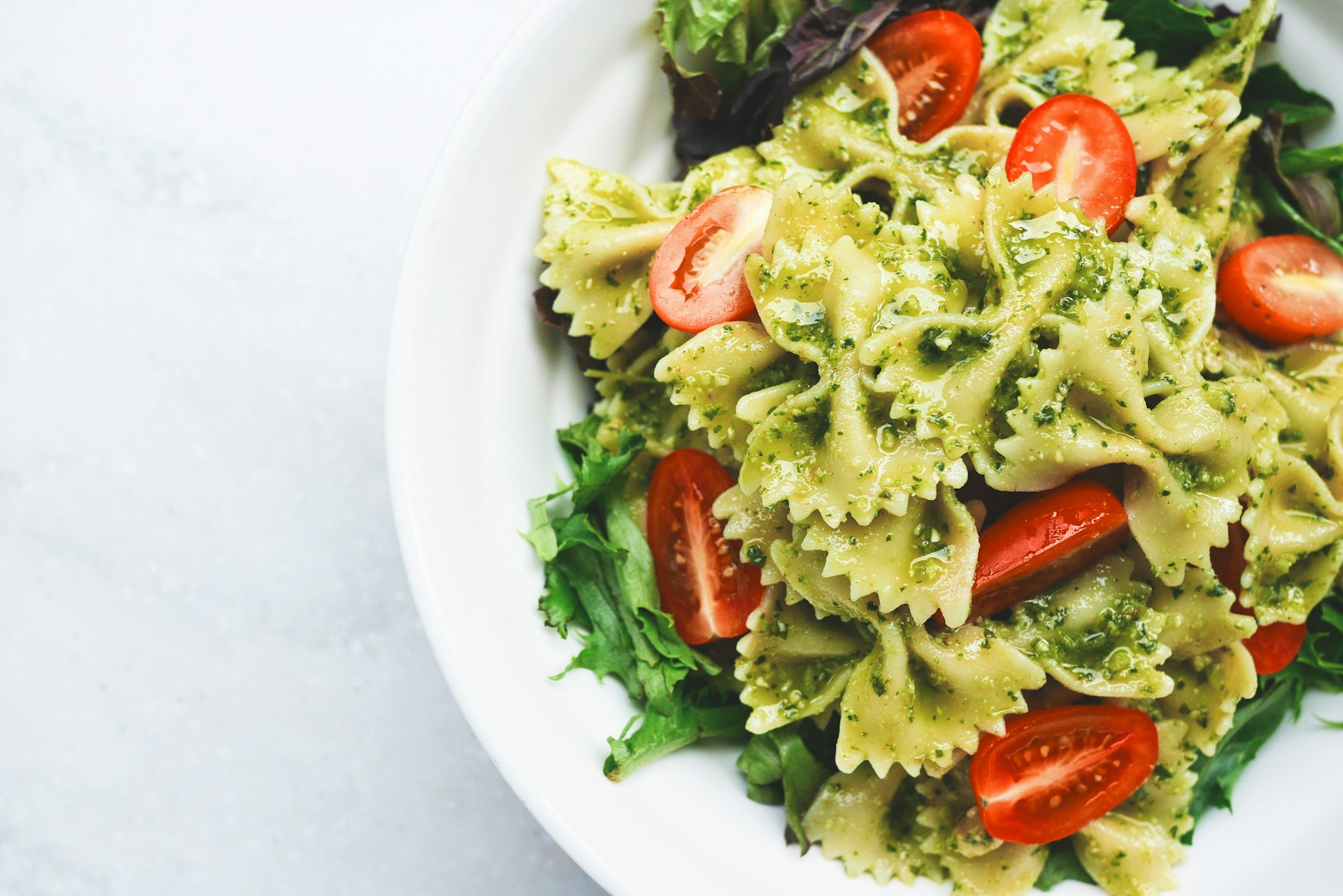 A bowl of farfalle pasta with pesto sauce, mixed greens, and cherry tomato halves.