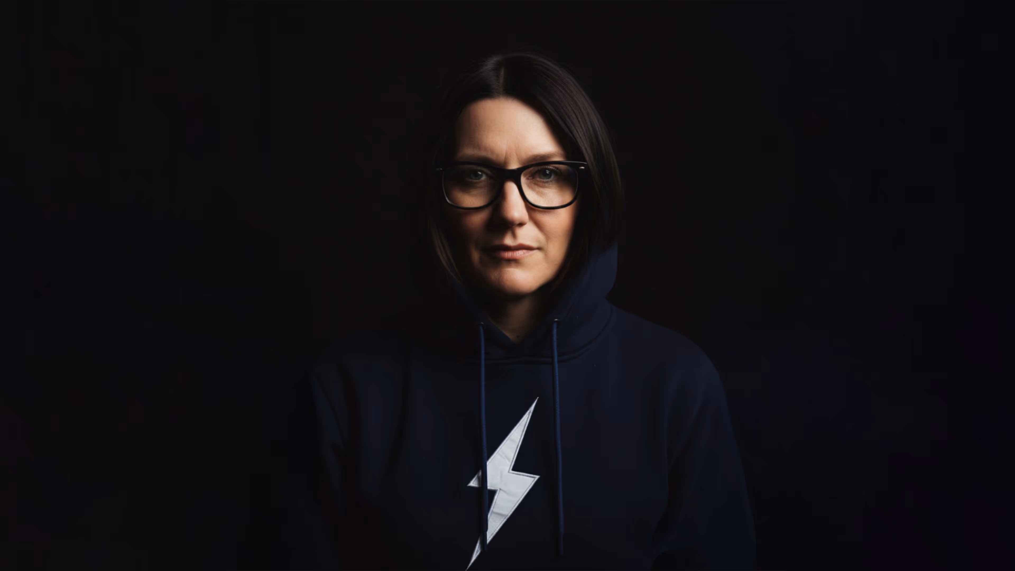 A dramatic, low-key headshot of a woman, Katie Swanson, looking directly at the camera. She is wearing black-rimmed glasses and a dark blue or black hoodie with a prominent white lightning bolt graphic on the chest. The background is solid black, creating a high-contrast and intense portrait.