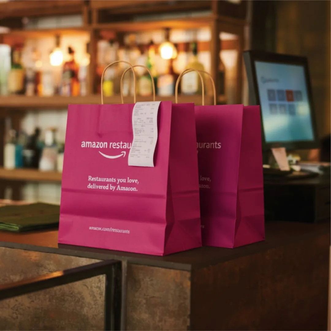 Two magenta Amazon Restaurants delivery bags with receipt on counter in restaurant setting.