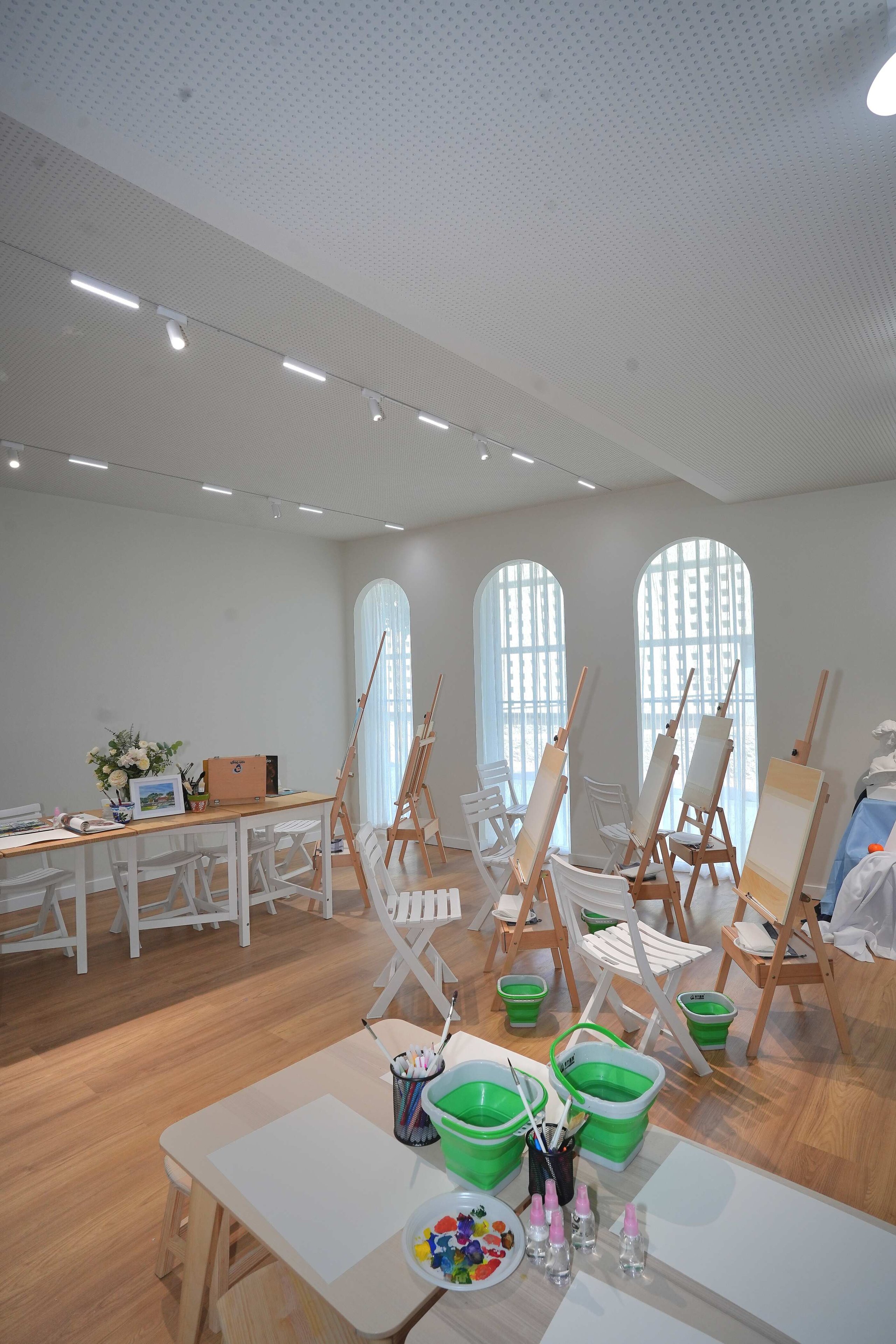 Aula de pintura con caballetes de madera, sillas blancas, cubetas verdes, pinceles y paletas de colores sobre mesas de madera clara en un espacio luminoso con ventanas arqueadas.
