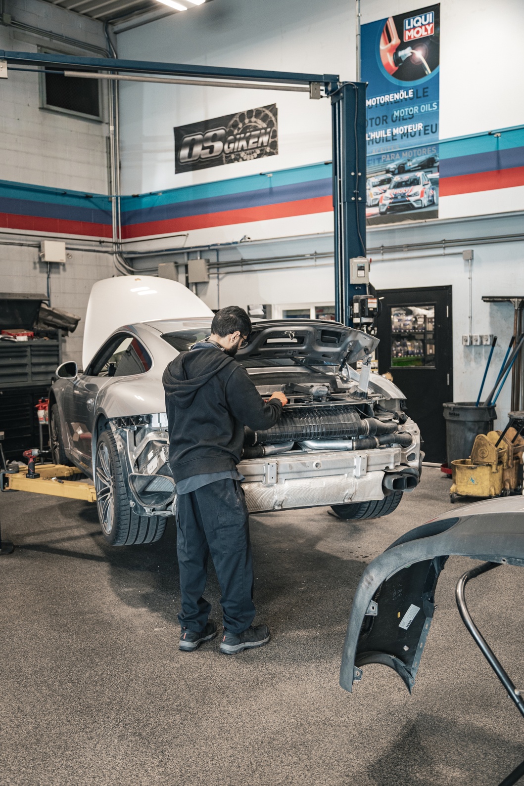 Mechanic working on the exposed engine of a silver sports car inside an auto repair shop.