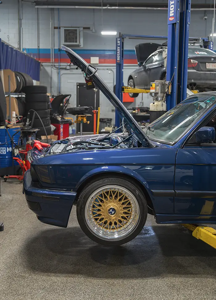 Blue BMW car with hood open showing engine in a garage with car lifts and tires.