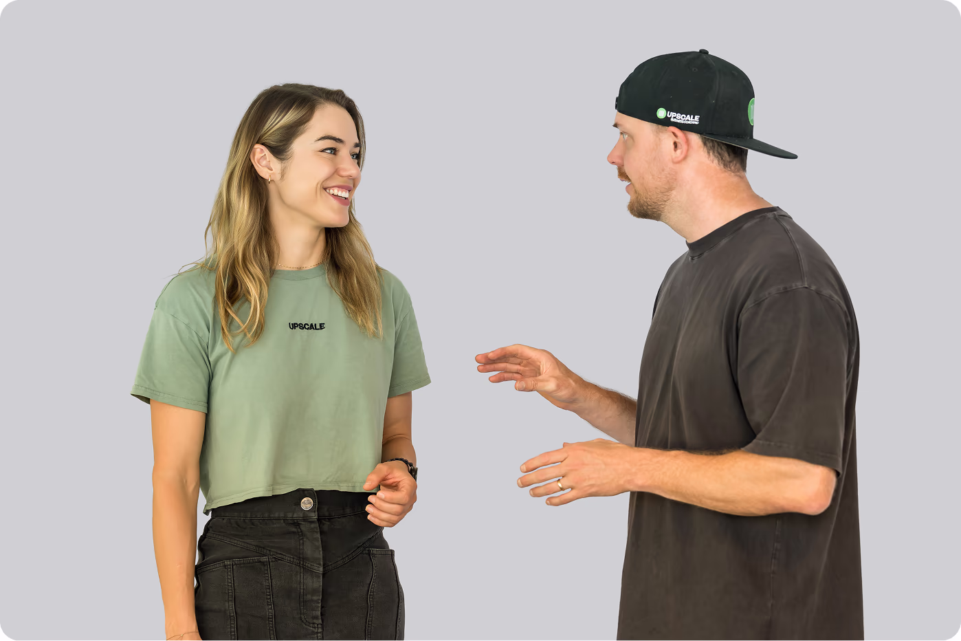 Smiling woman in green shirt talking with man in black shirt and cap against gray background.