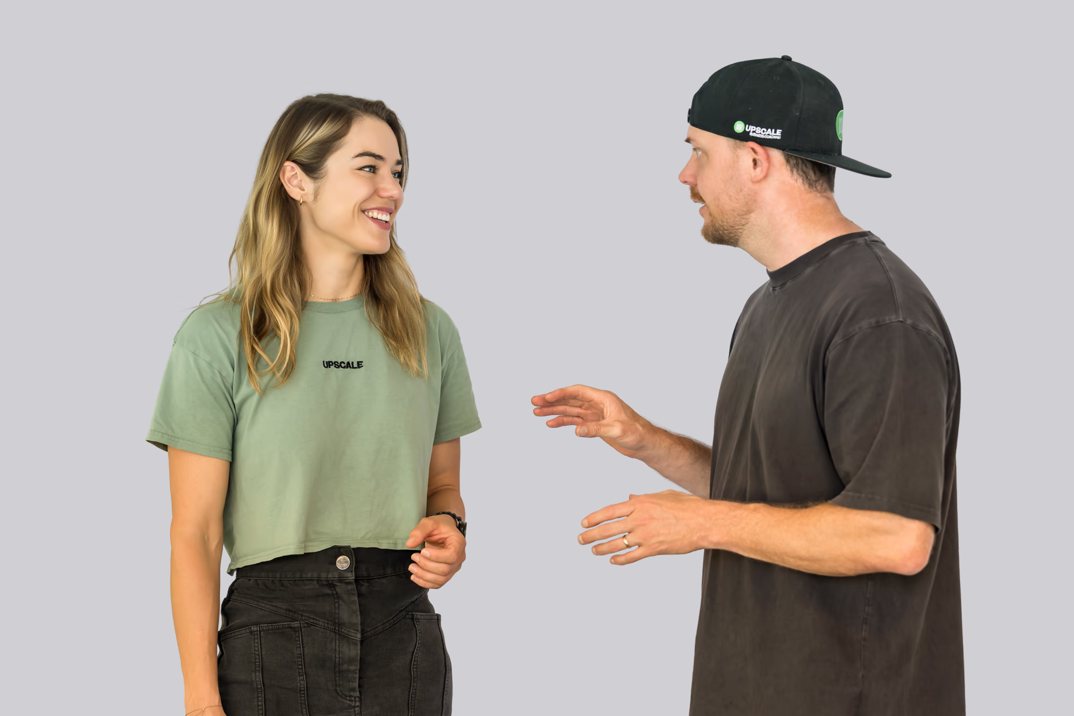 A man and woman in casual clothes facing each other and talking against a plain background.