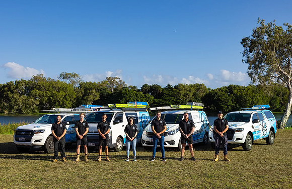 Seven people in black shirts standing in front of five service vehicles with ladders on a grassy field.