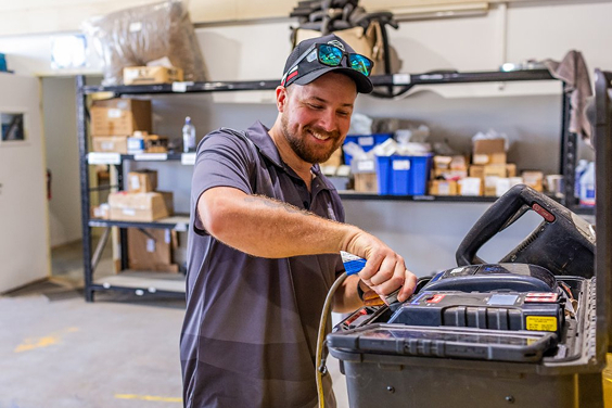 Smiling man in cap using a tape measure next to an open toolbox in a workshop with shelves.
