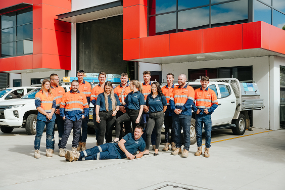 Group of workers in orange and blue uniforms posing outside a modern red and gray building.