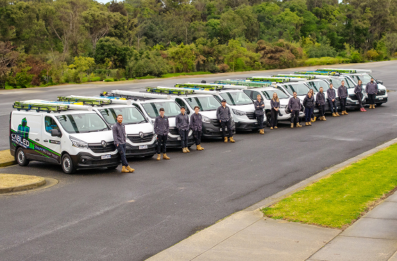 Team of workers standing beside a line of white company vans with ladders on top in a paved area.