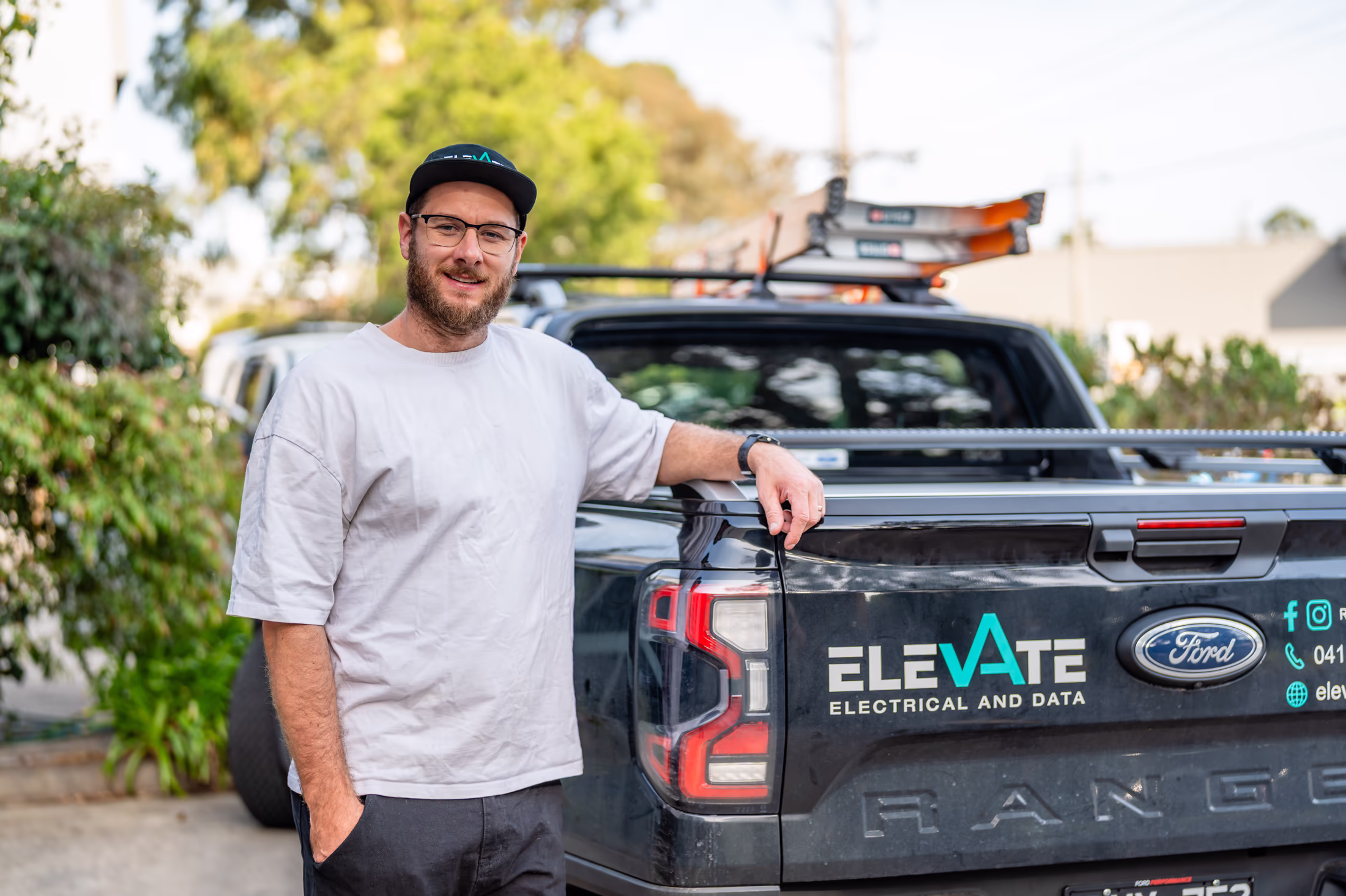 Man wearing glasses, a black cap, white t-shirt, and black pants stands smiling with one hand in his pocket and the other resting on the tailgate of a Ford Ranger truck with a logo and text reading 'ELEVATE Electrical and Data'.