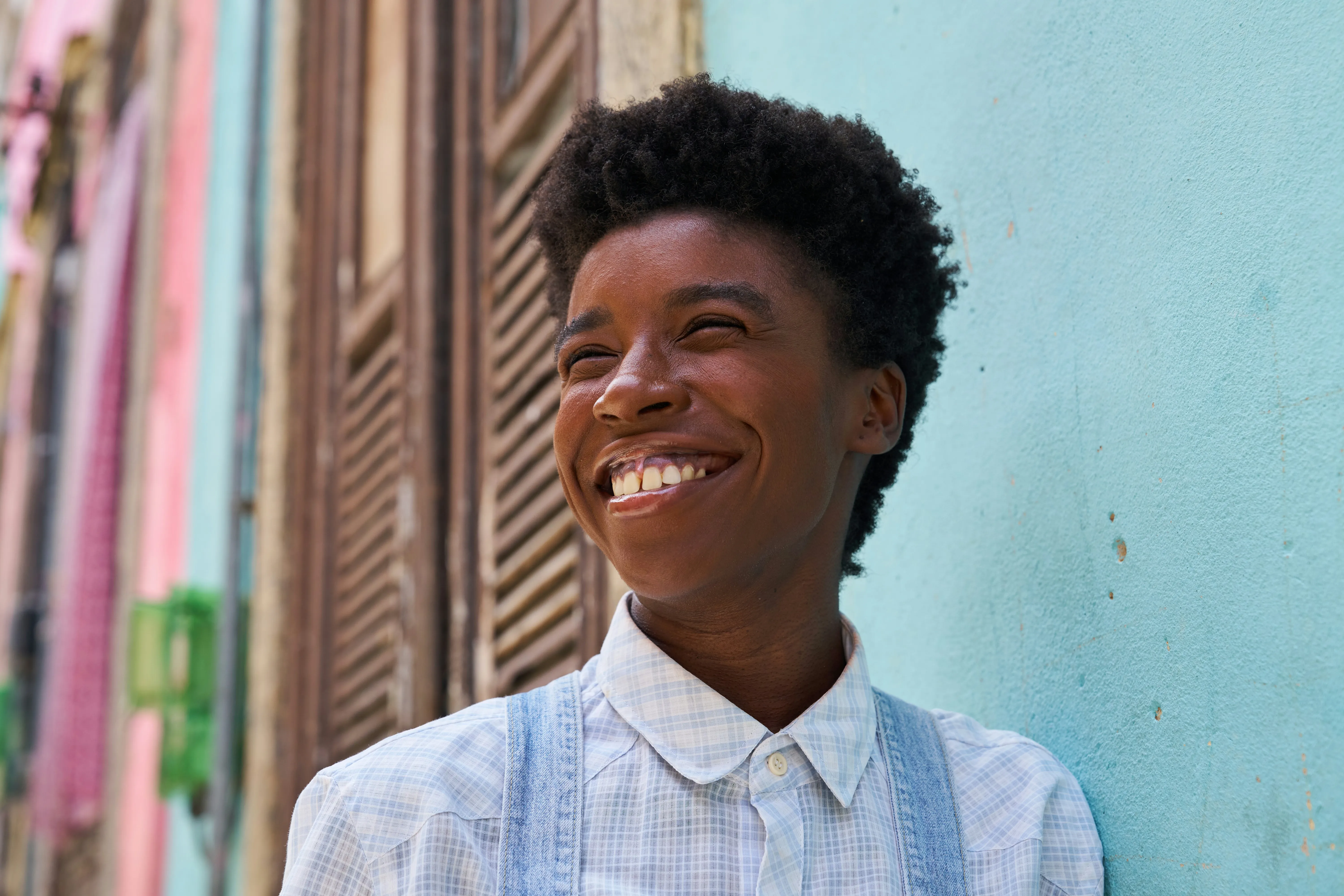 Smiling person with short curly hair wearing a checkered shirt and denim overalls, standing against a light blue wall with colorful buildings in the background.