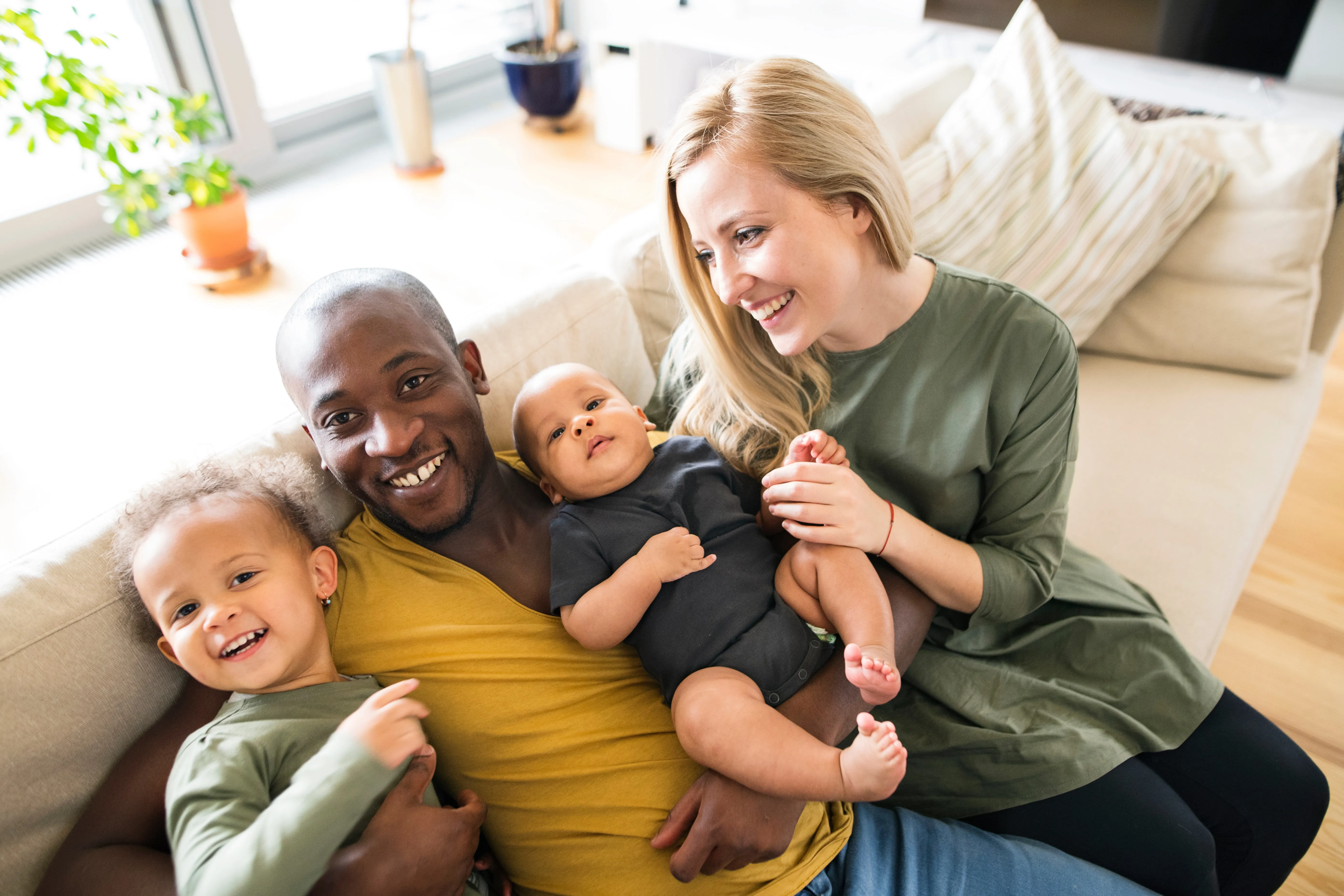 Happy mixed-race family of four with a father, mother, toddler, and baby sitting closely on a couch in a bright living room.